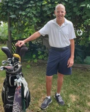 Man posing with golf clubs on grass, smiling, wearing a white shirt and shorts, green background.