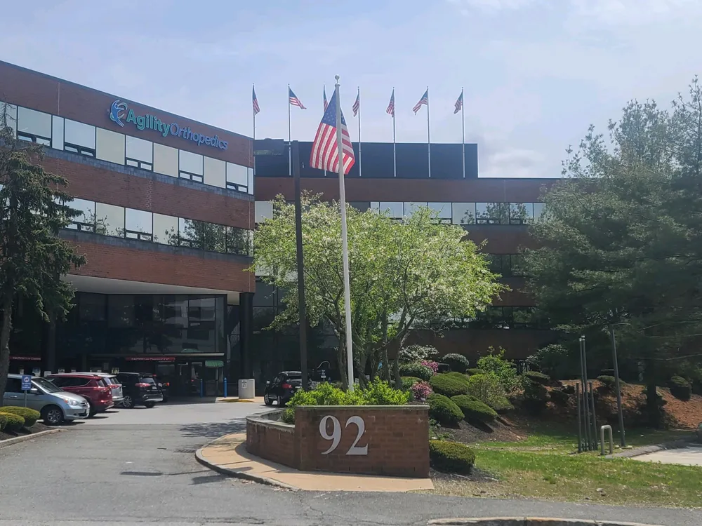 outdoor-stoneham-img Orthopedic clinic with American flag and number 92 on entrance sign, surrounded by trees and parked cars.