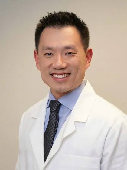 Smiling doctor in a white coat, light blue shirt, and dark tie against a neutral background.
